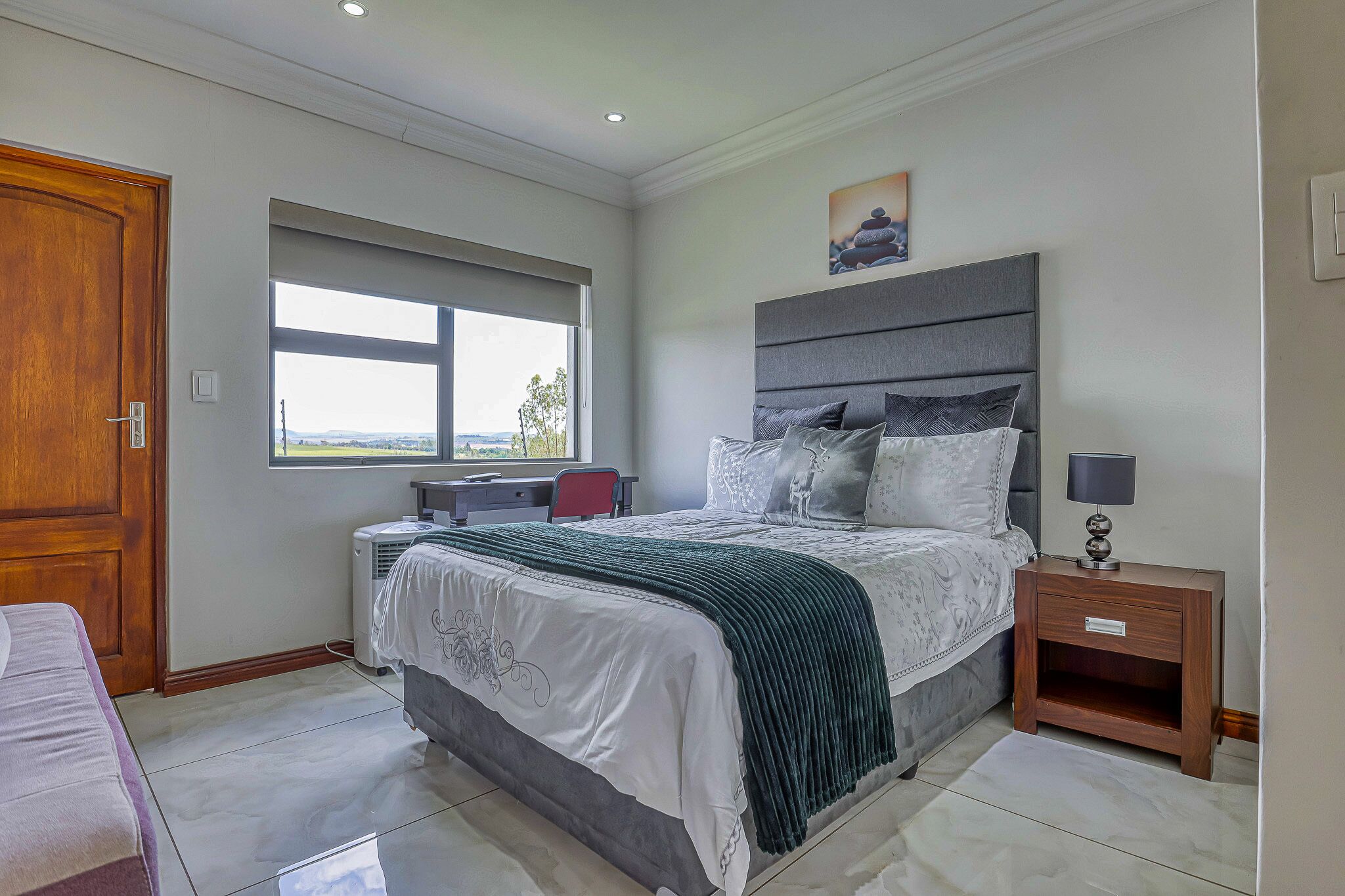 A bedroom with a large bed, a bedside table with a lamp, a window, and a desk with a chair.
