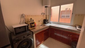 A compact kitchen with a washing machine, a coffee maker, and a window with a view. - Appartement Équipé et Climatisé (Marrakech)