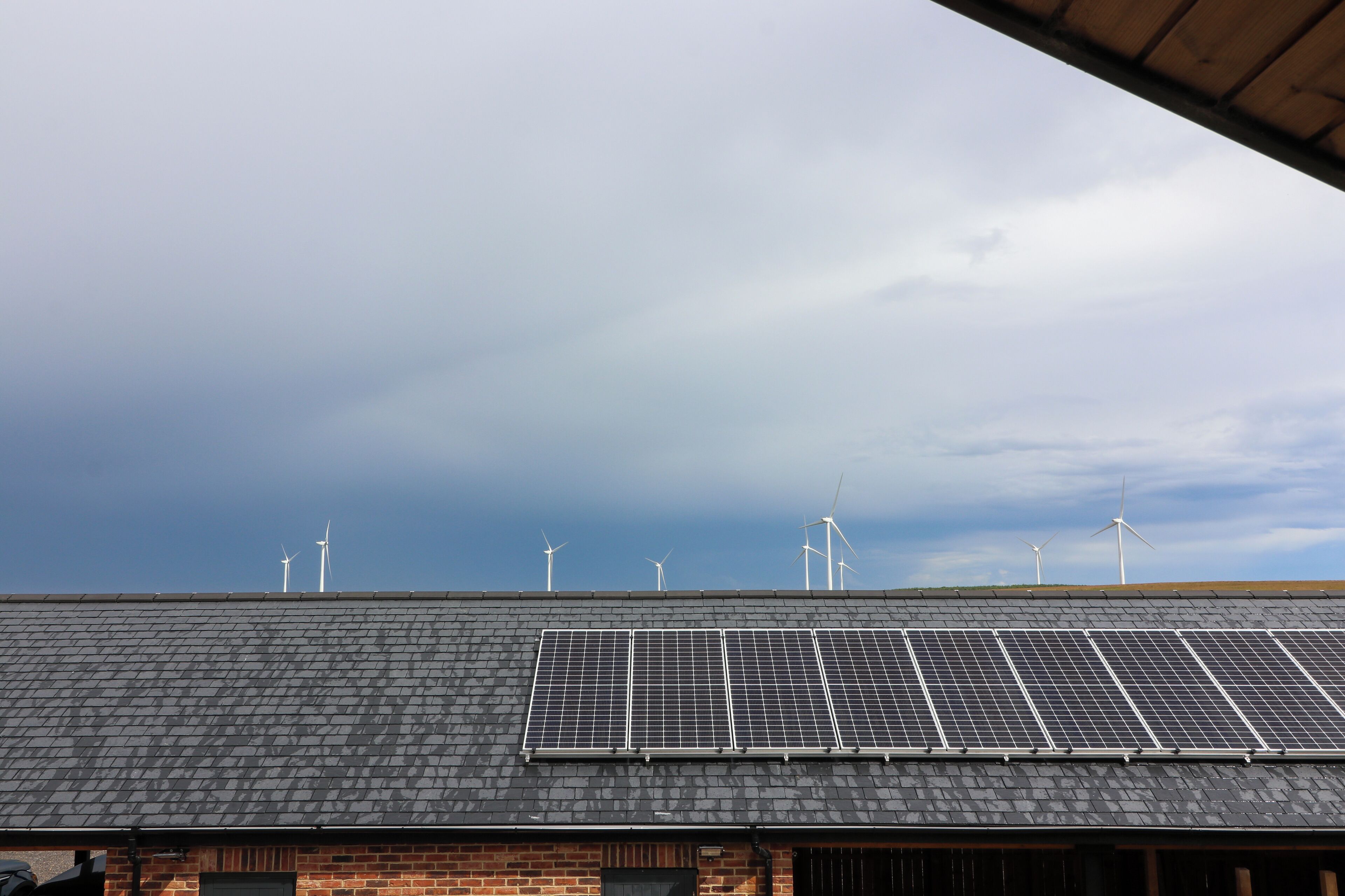 A building with a sloped roof, solar panels, and wind turbines in the background.