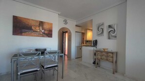 Een ruime woonkamer met een eettafel en stoelen, een keuken op de achtergrond en een gang naar een andere kamer.