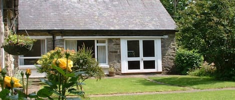 A stone house with a sloped roof, a front porch, and a small garden with yellow flowers.