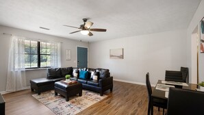 A living room with a black leather sofa, a coffee table, a dining table with chairs, a ceiling fan, and a window with sheer curtains. - Business-Ready 2 Bedroom Suite (Calhoun)