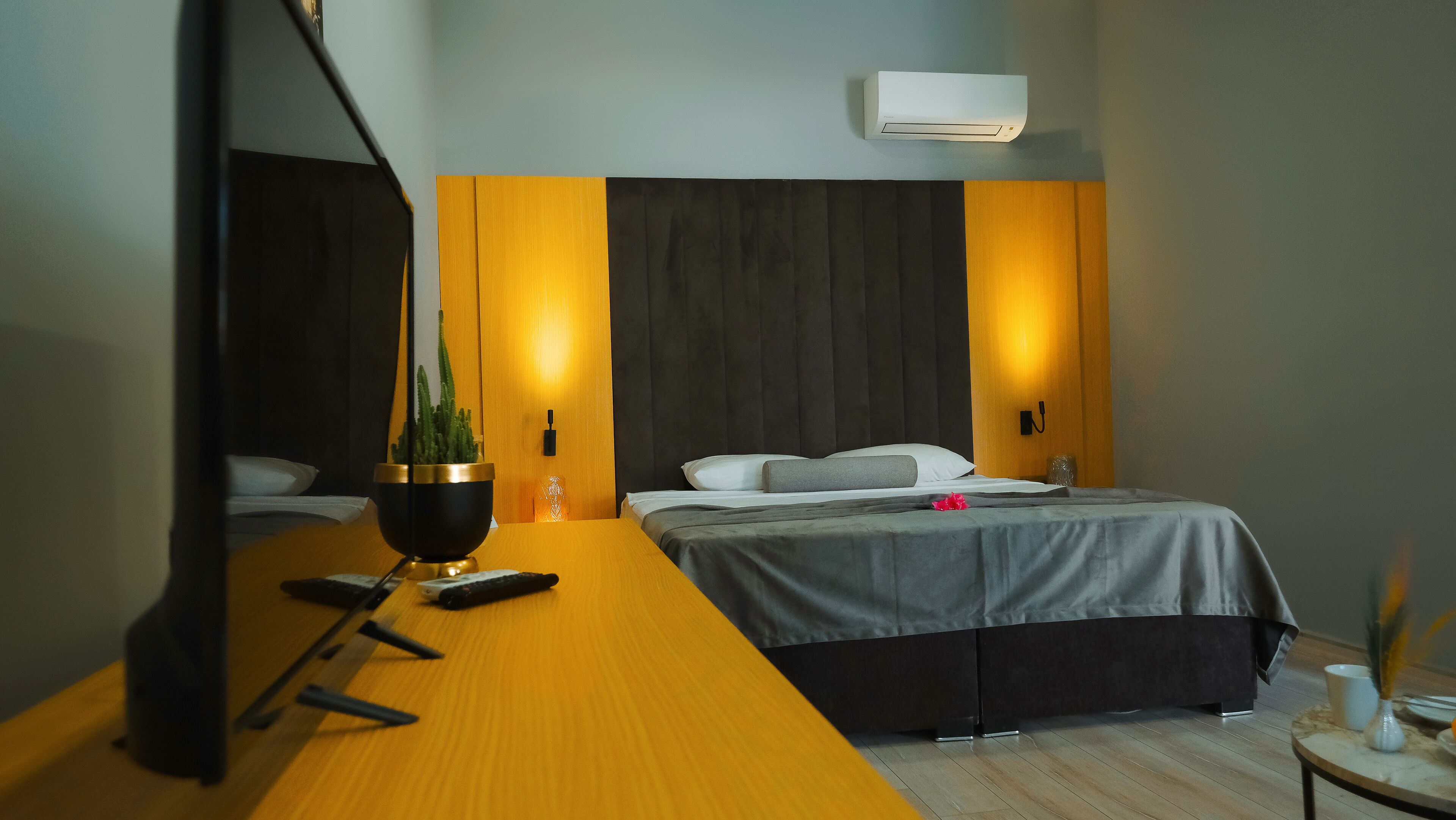 Una camera d'albergo moderna con una scrivania in legno, un letto con biancheria grigia, una pianta in vaso e un condizionatore d'aria a parete.