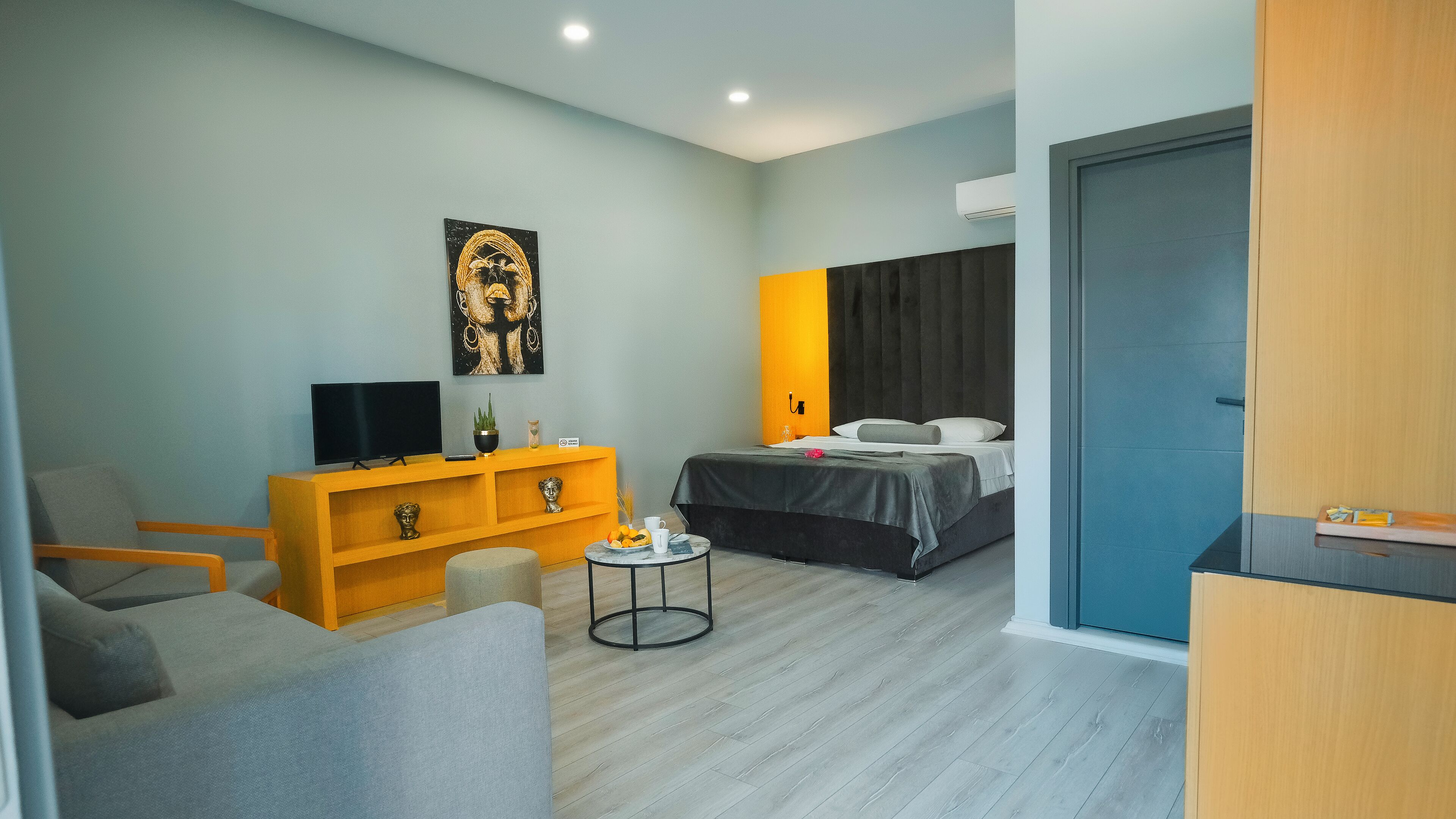 A modern hotel room with a sofa, a TV on a wooden console, a bed with a dark headboard, and a small round table with a plant.