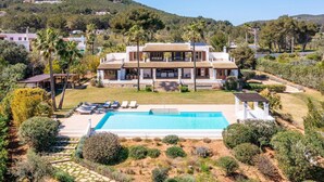 Une grande maison à deux étages avec une piscine, entourée d’une végétation luxuriante et avec des collines en arrière-plan.