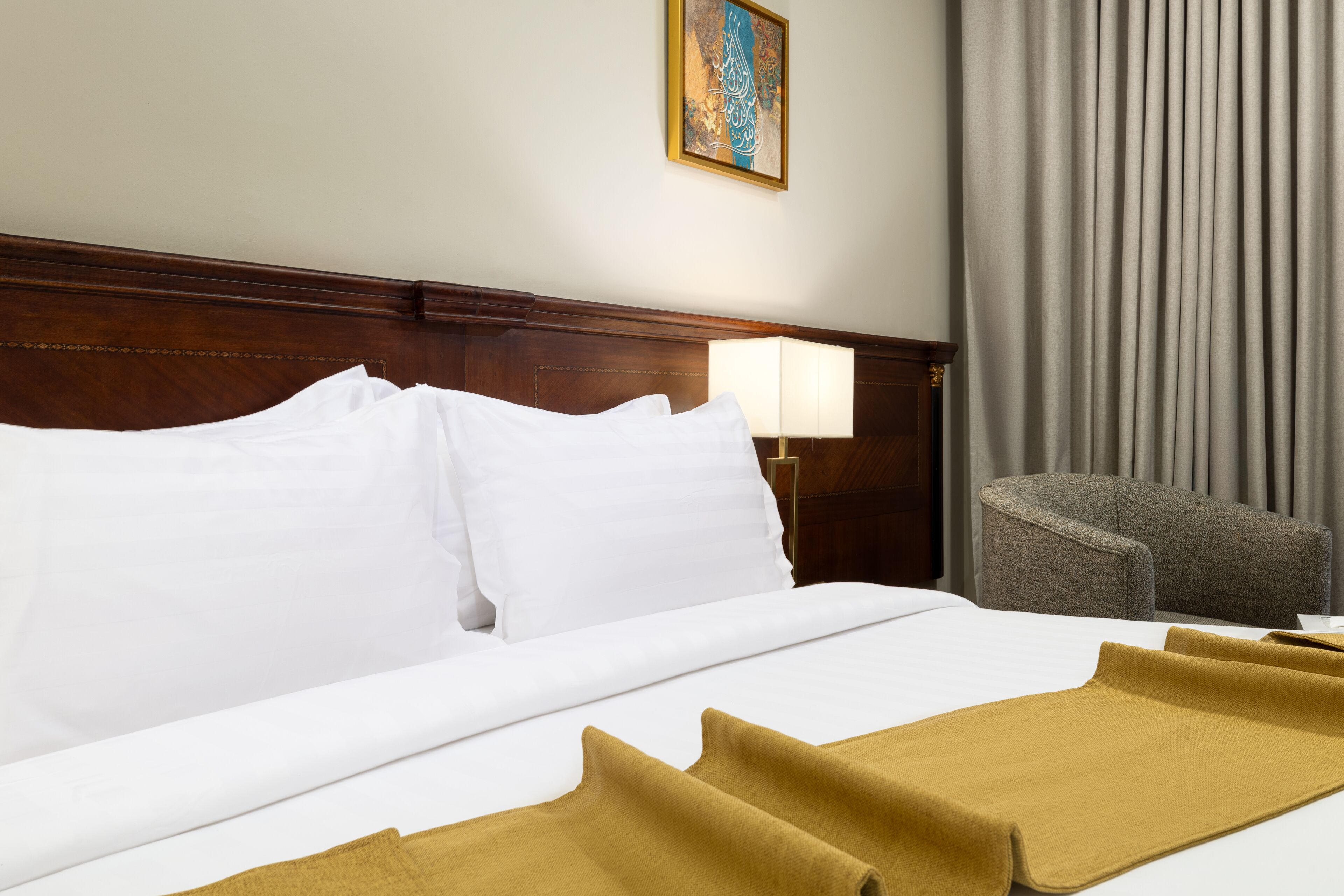 A hotel room with a wooden headboard, a bed with white linens, a grey armchair, and a lamp on a bedside table.