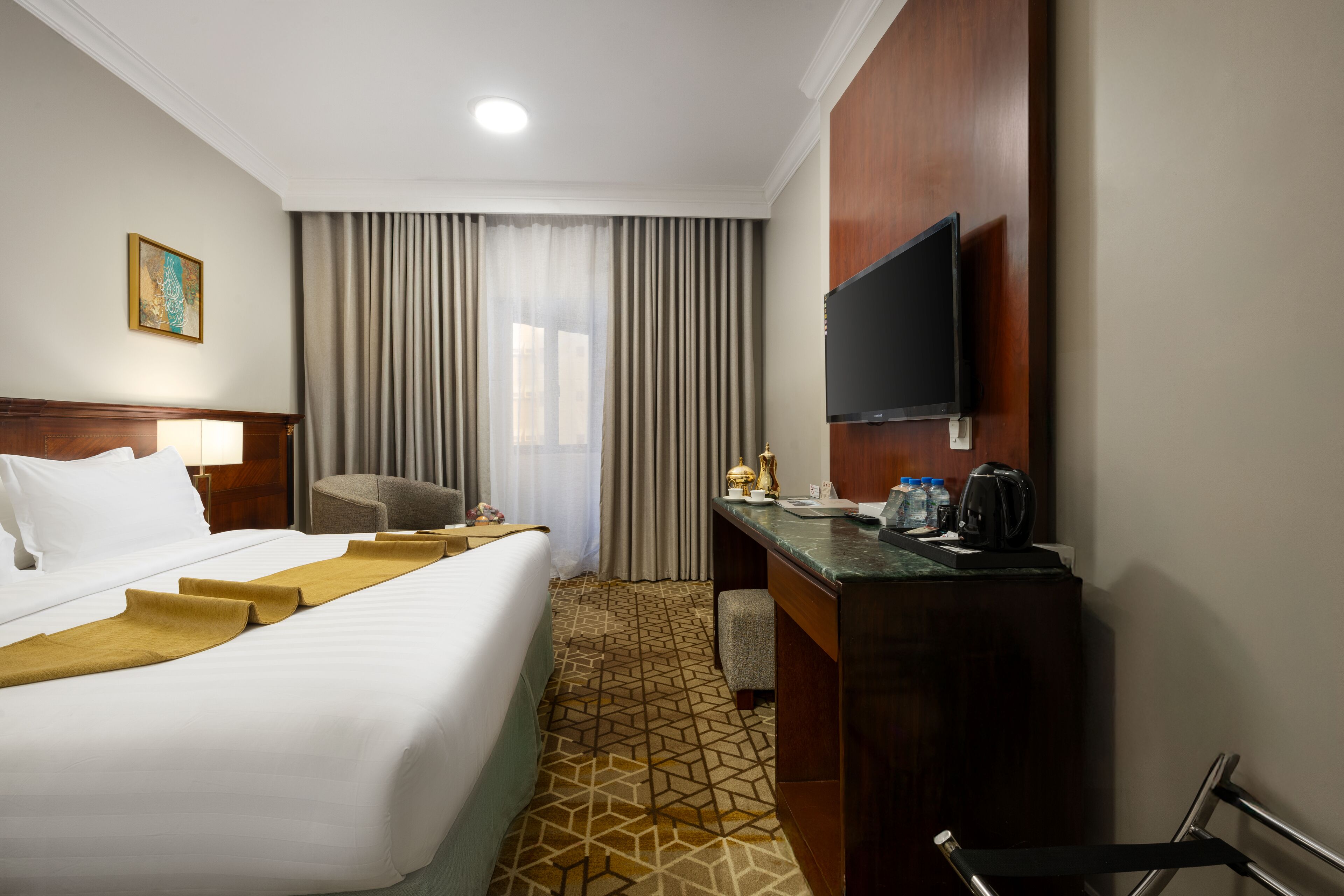 A hotel room with a large bed, a desk with a flat-screen TV, a chair, a desk lamp, and a coffee maker.