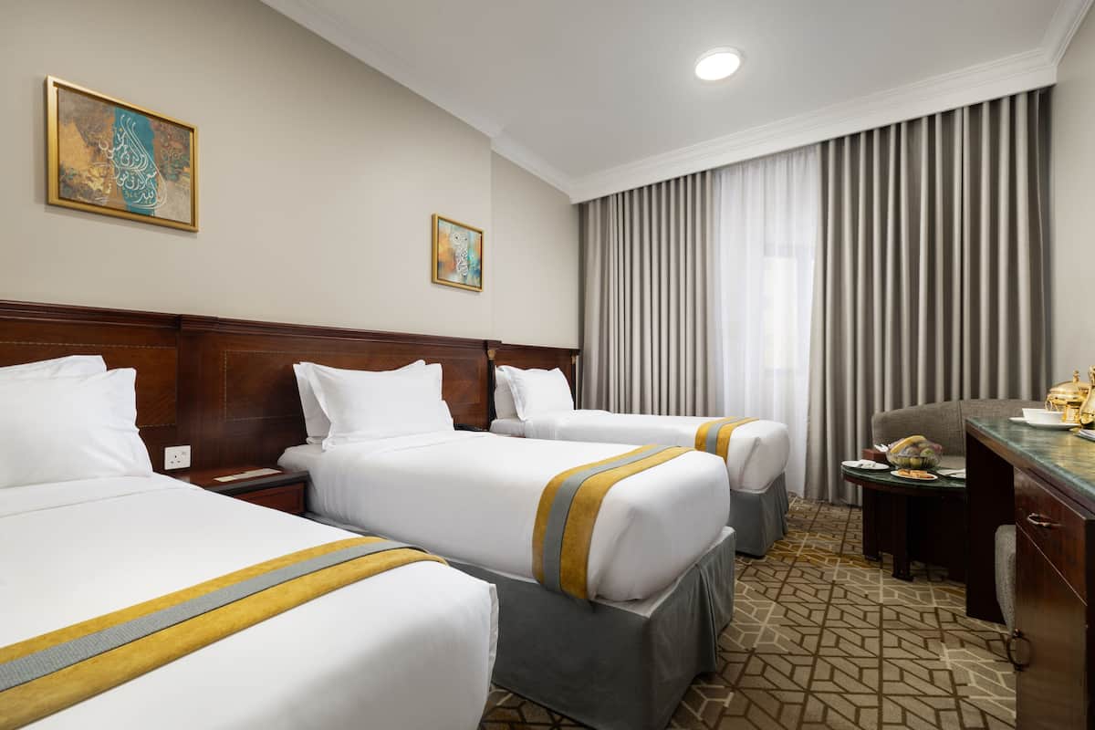 A hotel room with two beds, a desk, and a chair. There are paintings on the wall and a window with curtains.