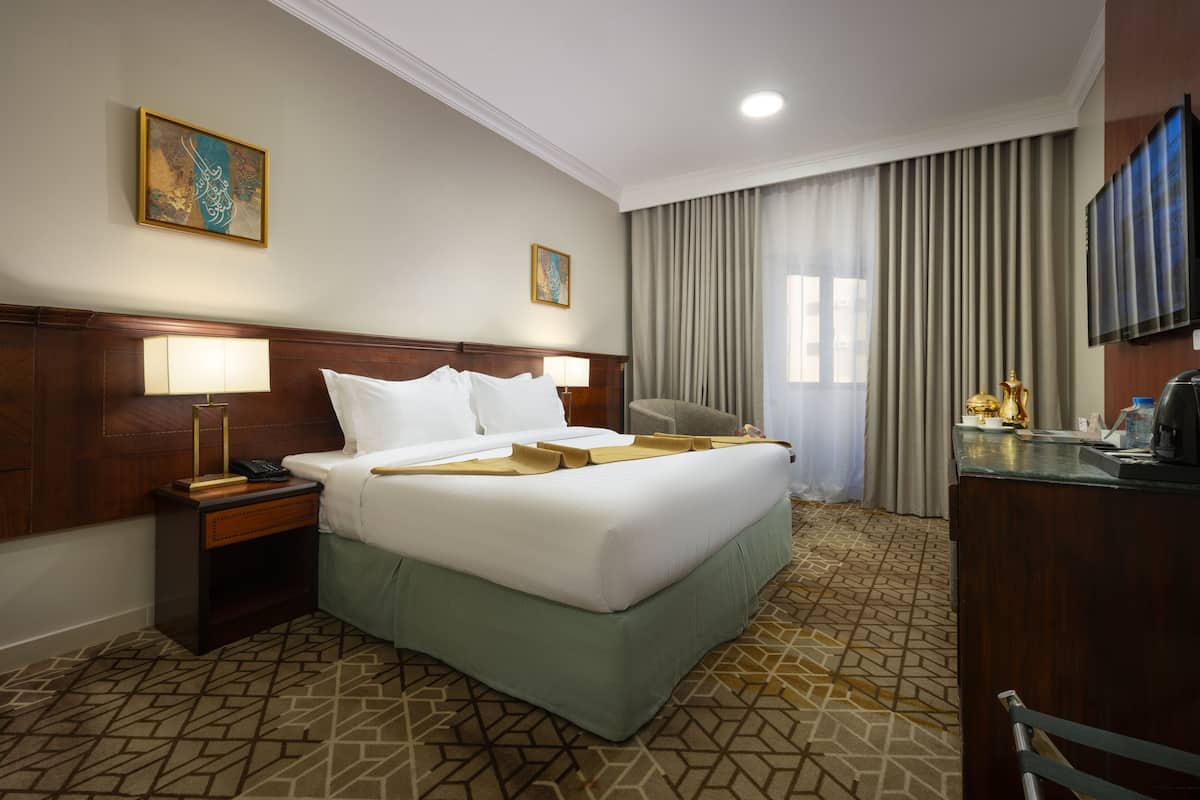 A hotel room with a large bed, a desk, a chair, a lamp, a telephone, and a television.