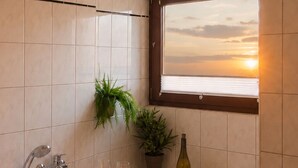 A bathroom with a bathtub filled with bubbles, a window with a sunset view, a potted plant, and a bottle of wine on the side.