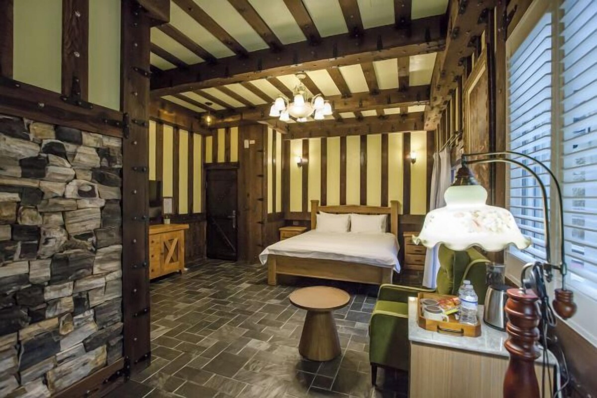 A bedroom with a stone wall, wooden beams, a bed, a nightstand, a lamp, and a small table.