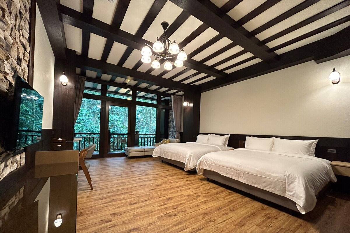A bedroom with two beds, wooden floors, a stone wall, and large windows overlooking a forest.