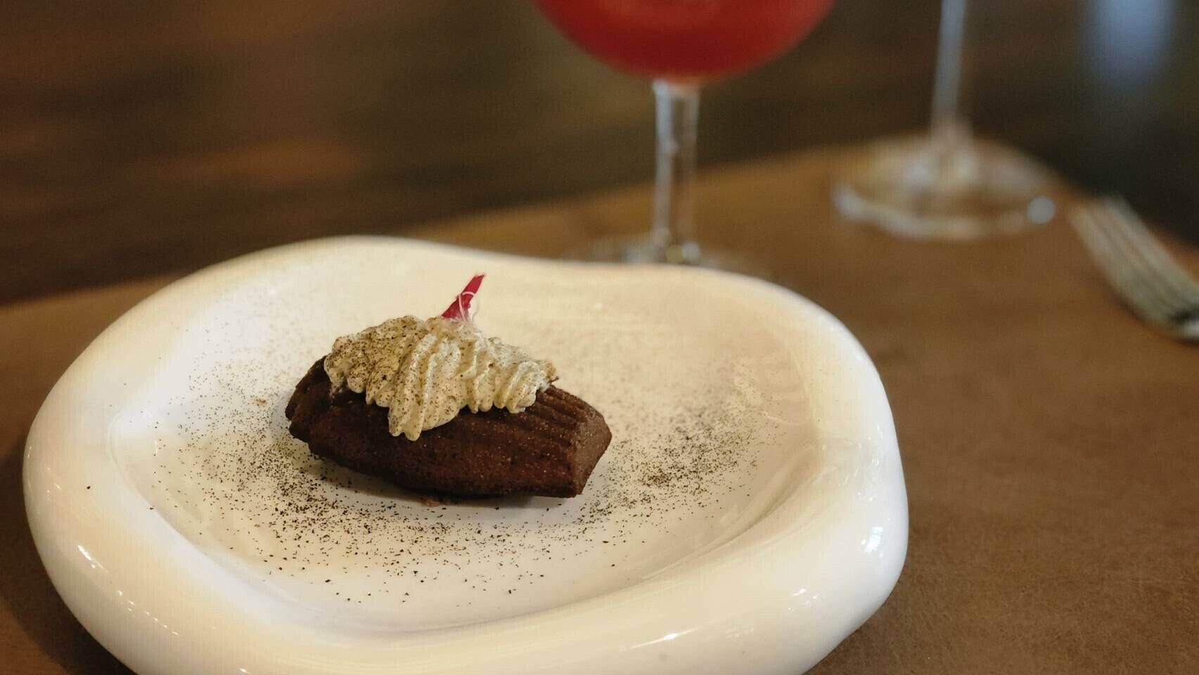 Dessert auf einem weißen Teller mit roter Garnitur, im Hintergrund ein Glas rotes Getränk und eine Gabel auf dem Tisch.