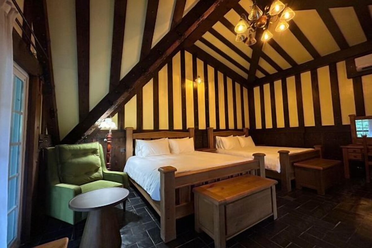 A room with two beds, a green armchair, a small round table, and a wooden bench.