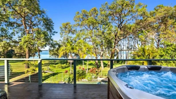 A hot tub on a wooden deck with a view of a lush garden and trees. - Blissful Lakefront Pool & Oasis (Lake Munmorah)