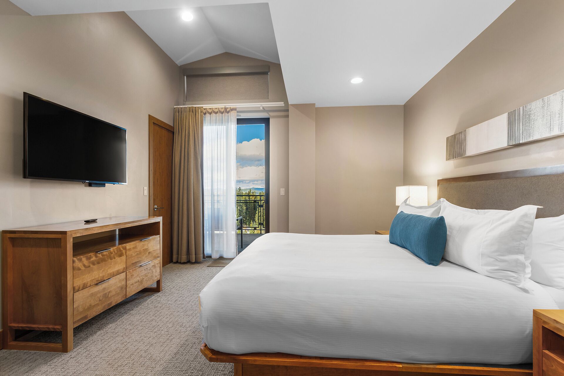 A hotel room with a large bed, a TV mounted on the wall, a wooden dresser, and a view of the outdoors through a window with curtains.