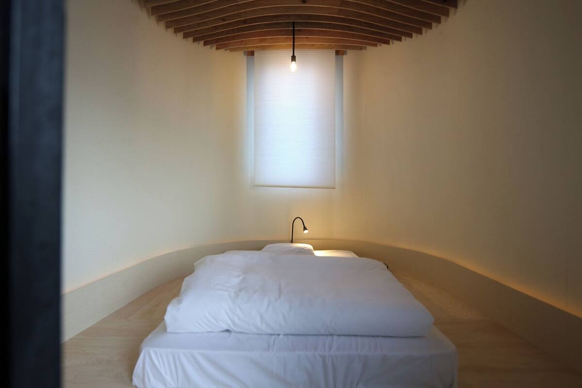 Un dormitorio minimalista con techo inclinado, una cama individual, una lámpara de noche y una ventana con persianas.