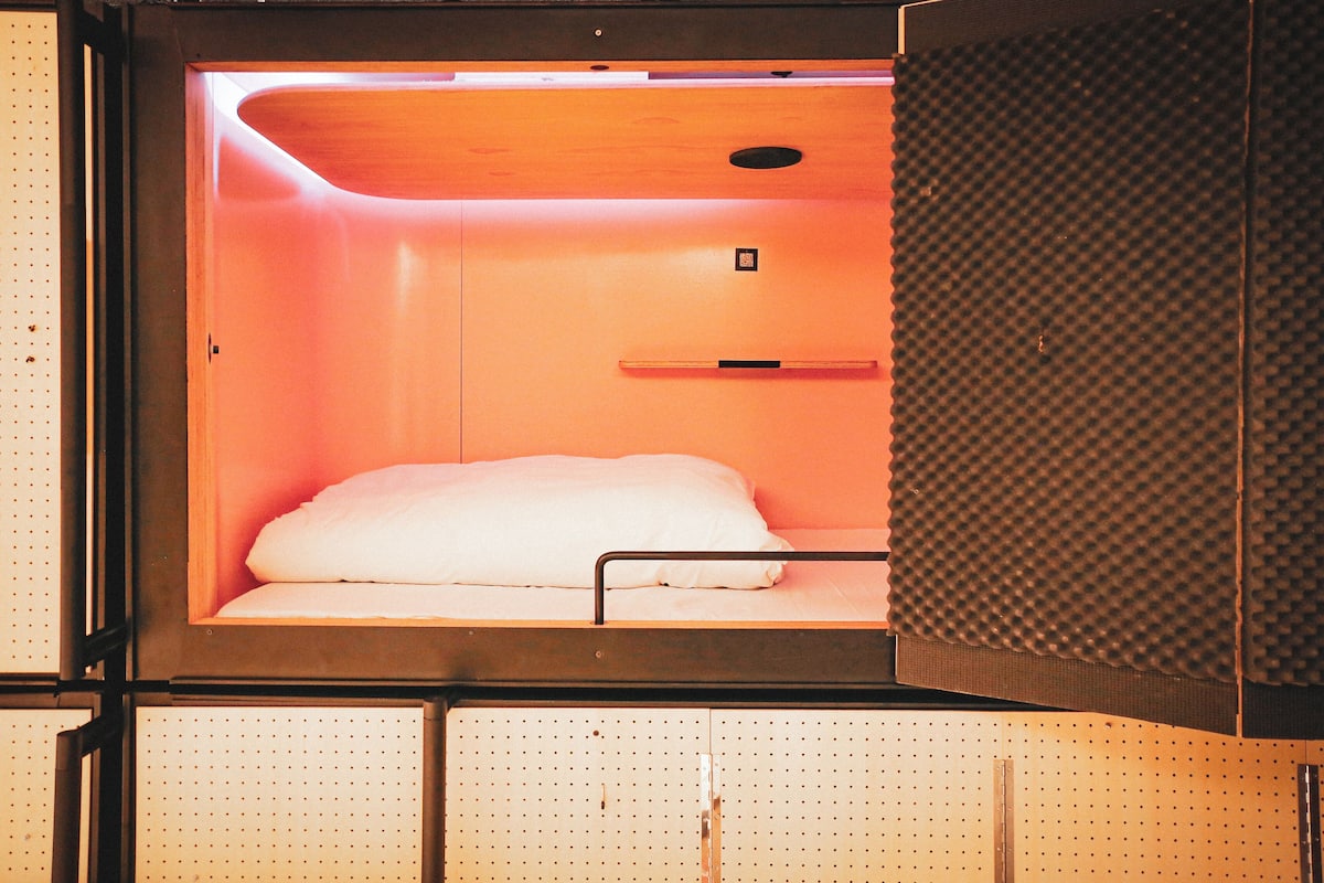 Una habitación compacta y moderna con una cama individual, un pequeño estante y un panel de control.