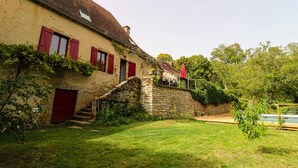 Une maison de campagne en pierre, avec des volets rouges, un escalier en pierre et une cour couverte d’herbe.