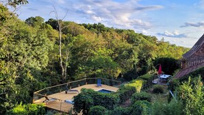 Une terrasse en bois avec une piscine, entourée d’une végétation luxuriante et agrémentée d’un parasol rouge.