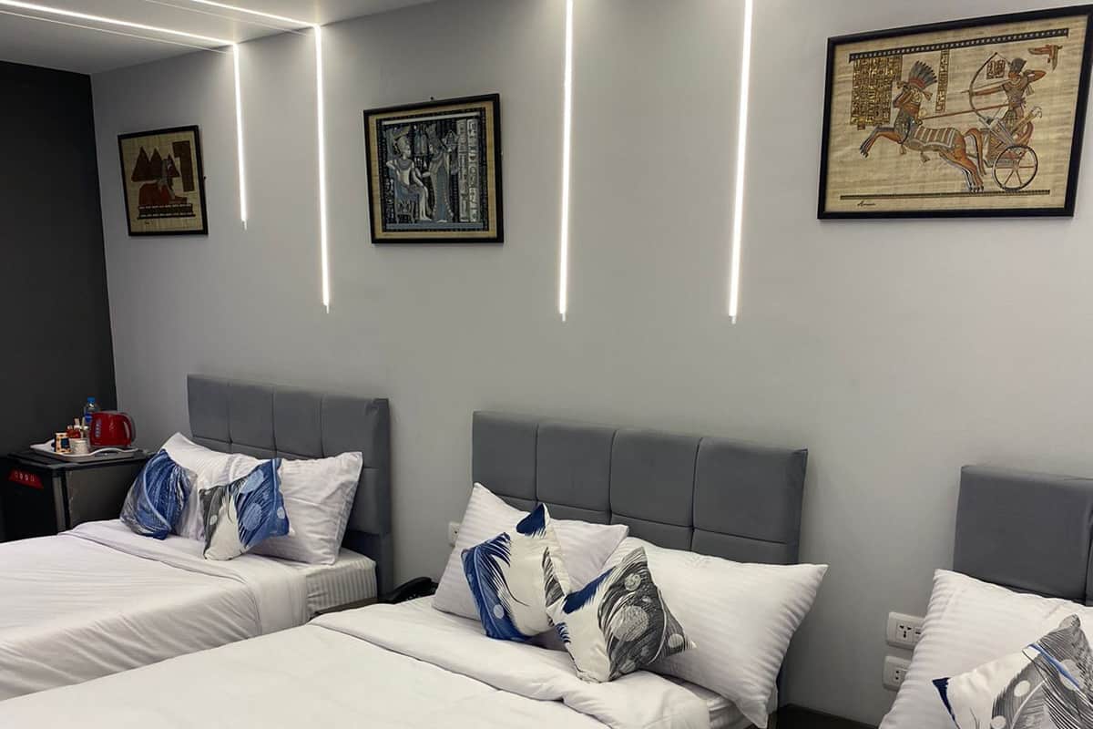 A hotel room with three beds, each with white bedding and decorative pillows. The room features modern lighting with linear LED strips on the ceiling.