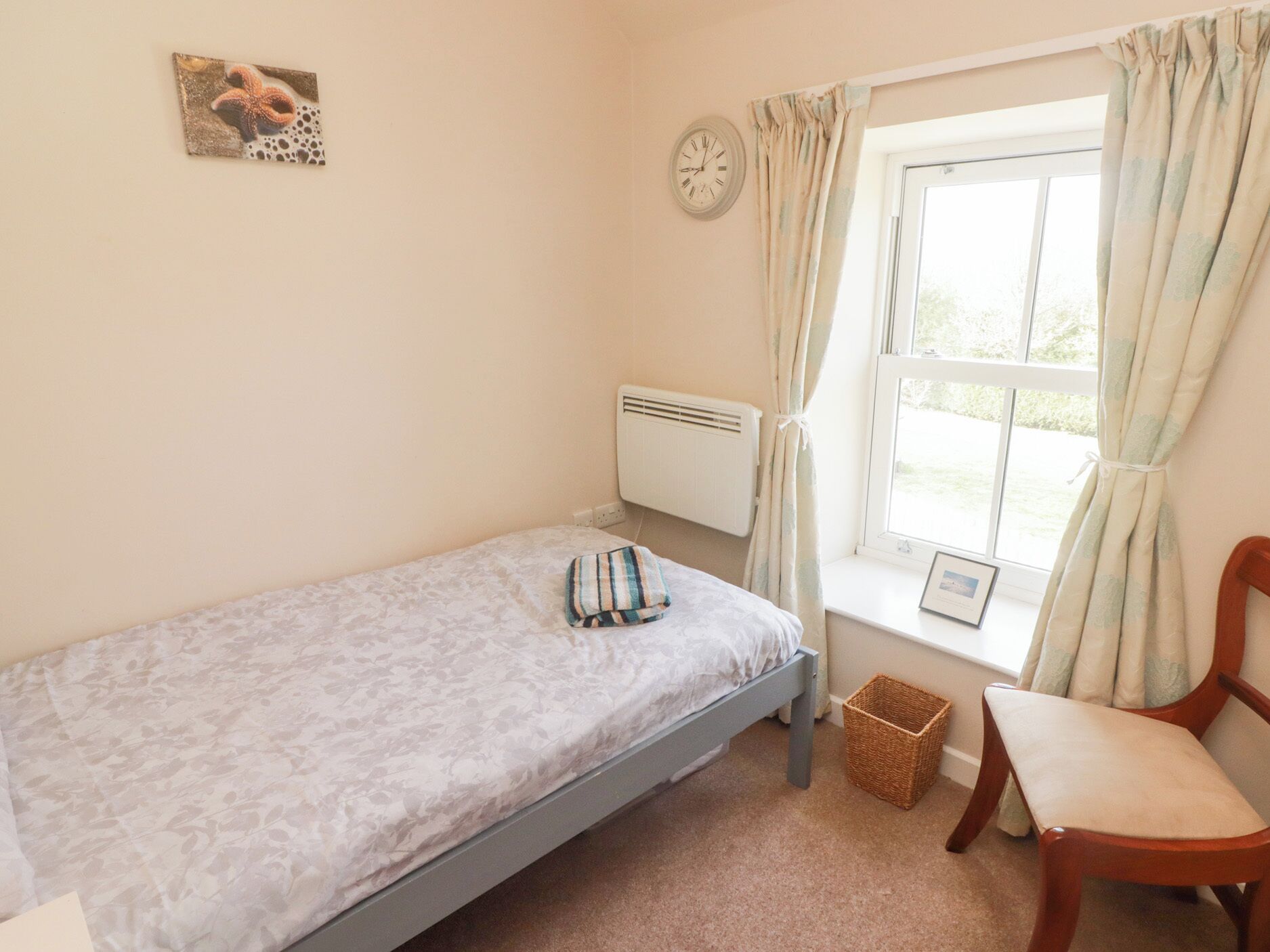 A small, single-bed room with a window, a chair, a clock, and a radiator.