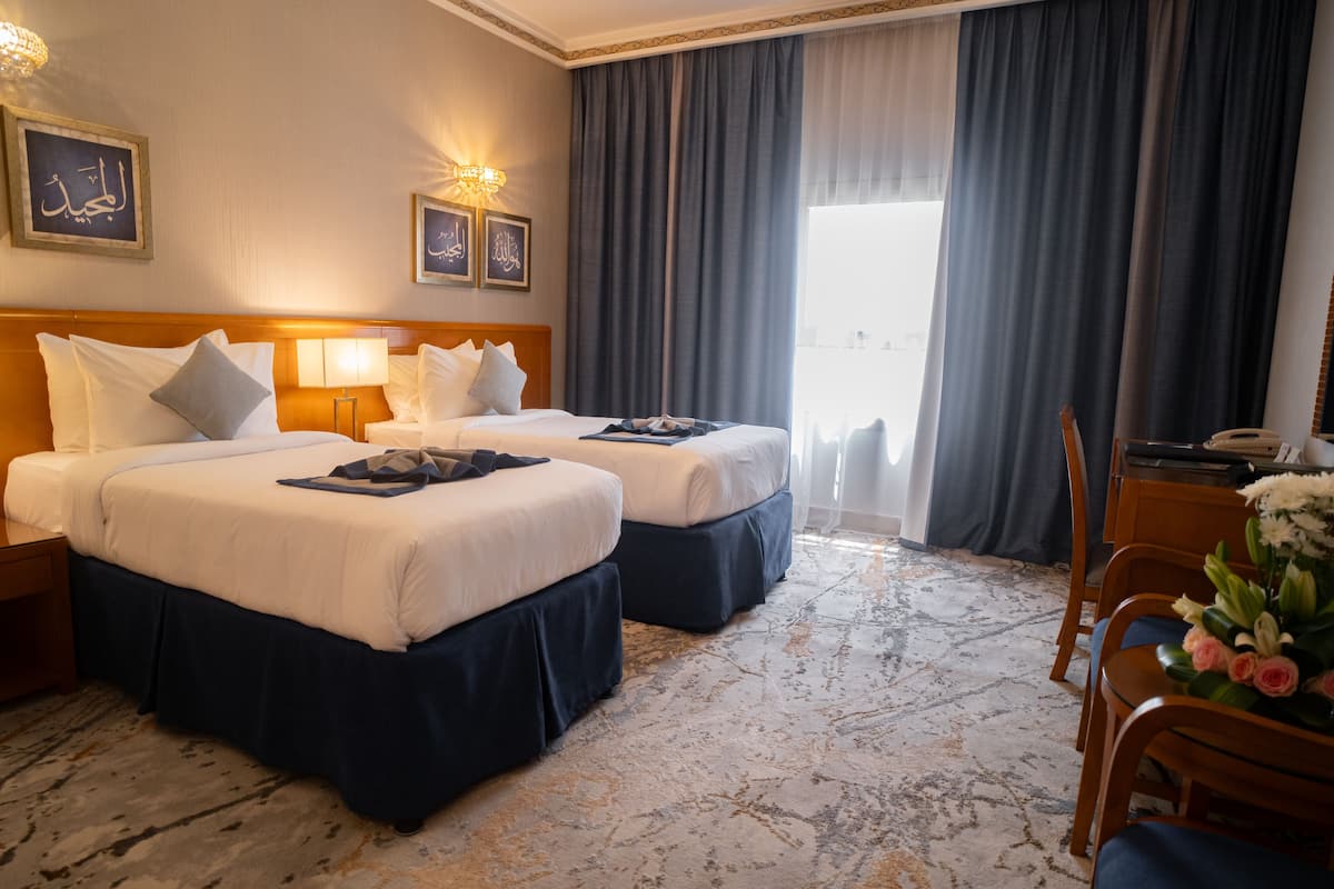 Ein Hotelzimmer mit zwei Betten, einem Schreibtisch, einem Stuhl und einem großen Fenster mit Vorhängen.