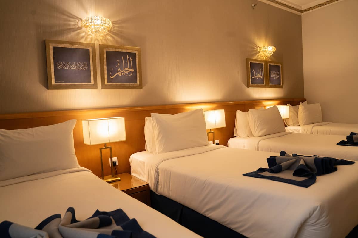 Ein Hotelzimmer mit vier Betten, jeweils mit weißer Bettwäsche und einem hölzernen Kopfteil. An den Wänden befinden sich Wandleuchten und gerahmte arabische Kalligraphie.