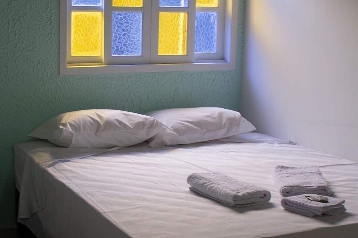 A small, neatly made bed with white linens and pillows, a window with a view of a yellow building, and a folded towel on the bed.