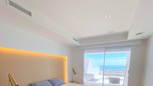 Un dormitorio moderno con una cama grande, una mesita de noche con lámpara y vistas al océano a través de un ventanal corredero.