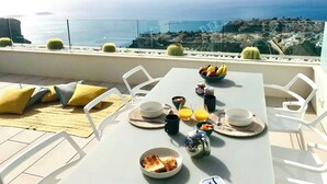 Una mesa de comedor con alimentos para el desayuno, sillas blancas y vistas al océano y al paisaje urbano.