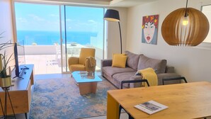 Una sala de estar moderna con sofá, mesa de centro y mesa de comedor. Una ventana grande ofrece vistas al océano.