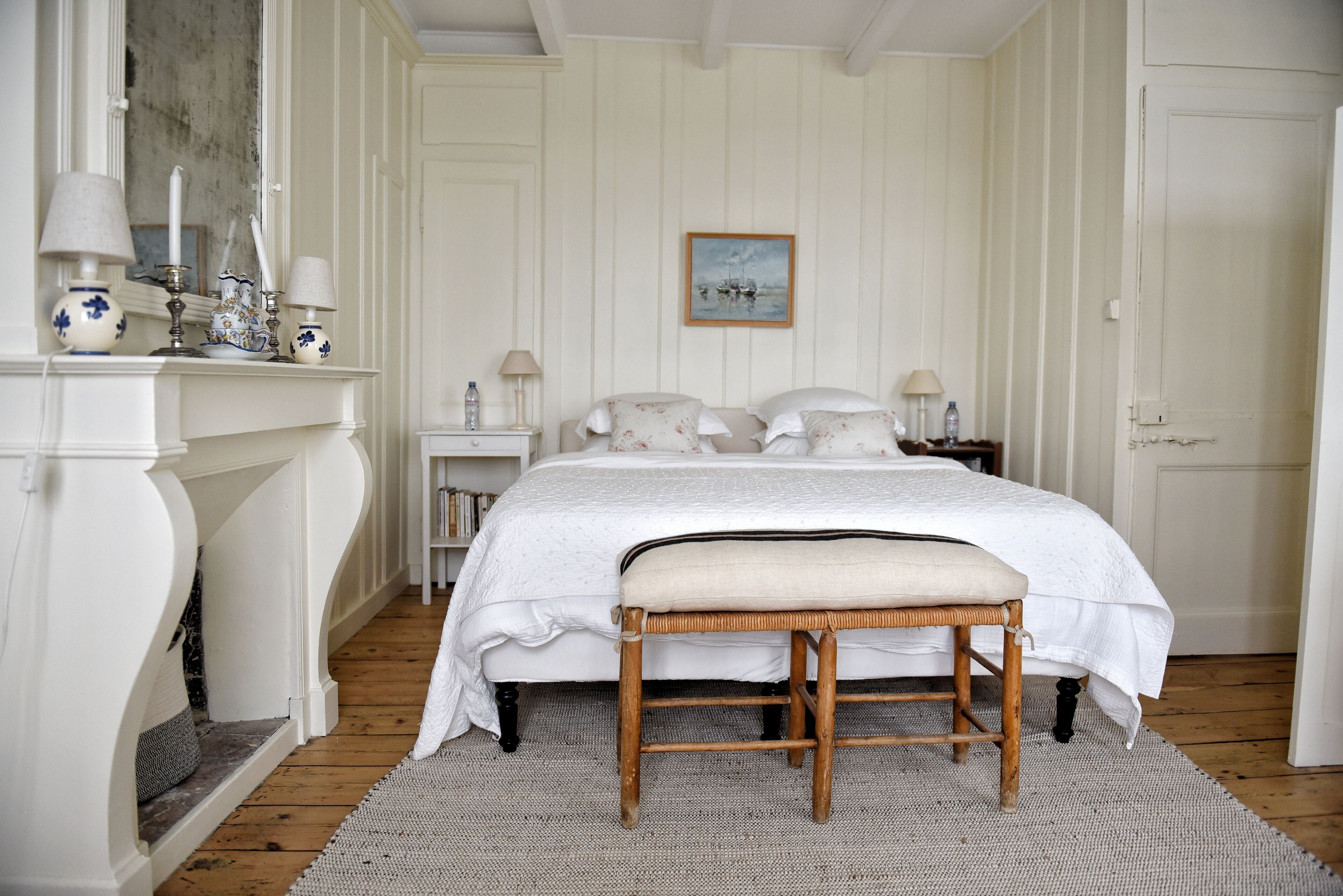 A bedroom with a bed, a bench, a fireplace, and a painting on the wall.