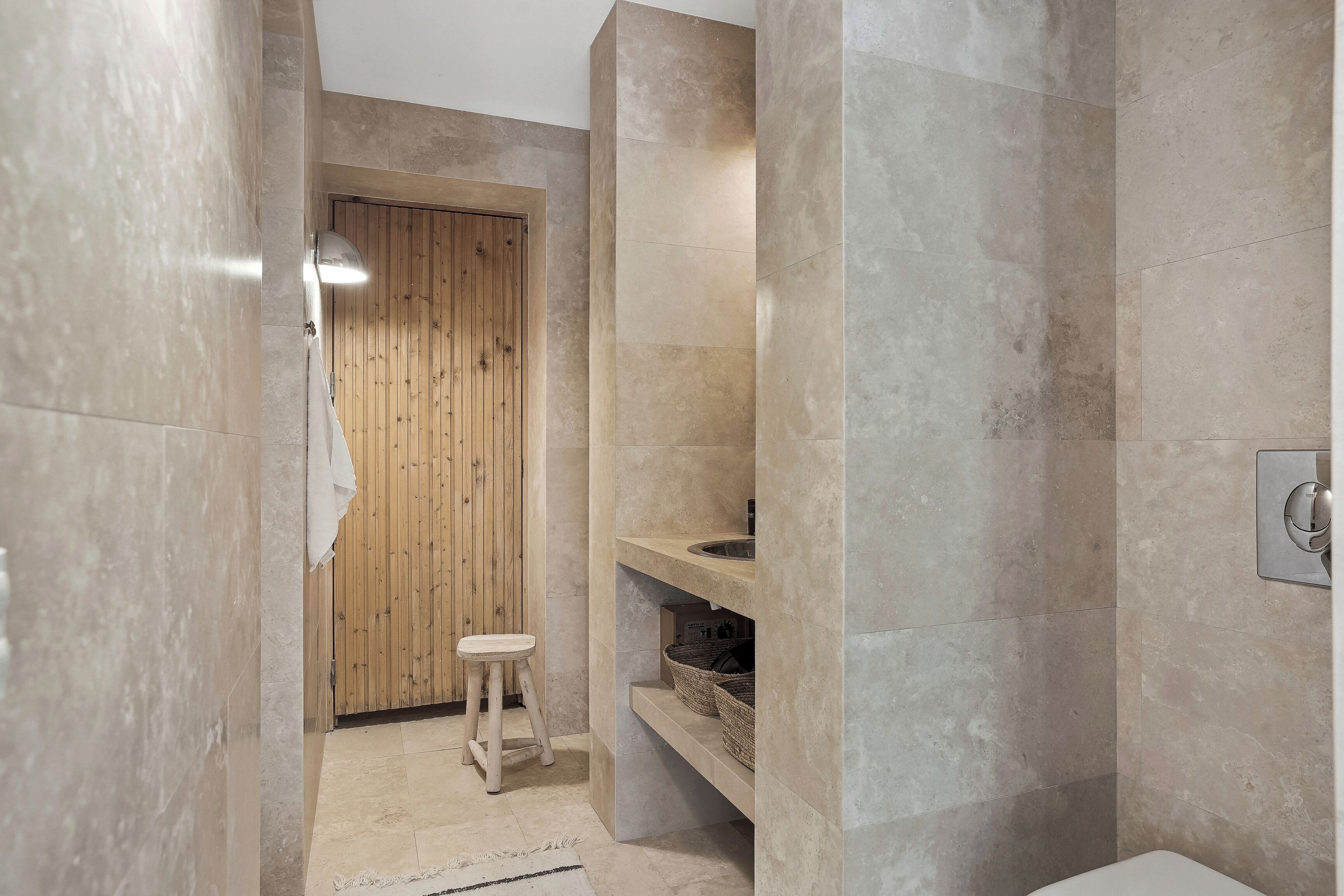 Un baño moderno con puerta de madera, un taburete pequeño, un lavamanos y una repisa con cestas.