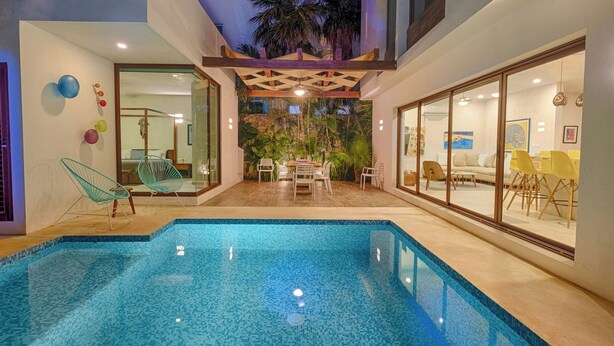 Un área de piscina moderna con un porche de cristal, una zona de comedor con sillas amarillas y un dormitorio con cama y sillón.