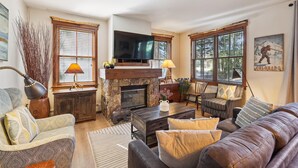 A cozy living room with a stone fireplace, comfortable seating, and large windows offering a view of the outdoors.