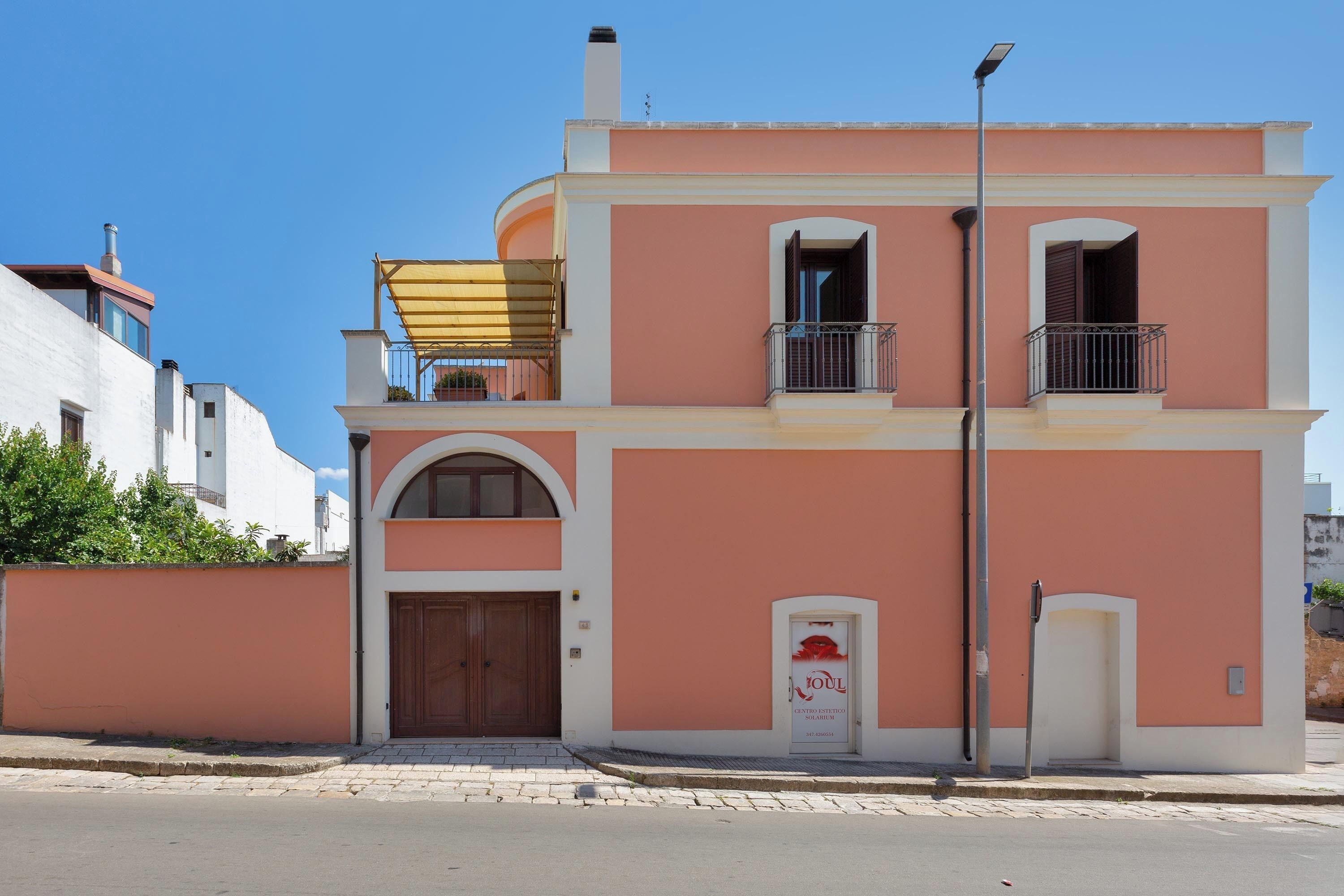 A two-story building with a pink facade, a wooden door, and a balcony with a metal railing.