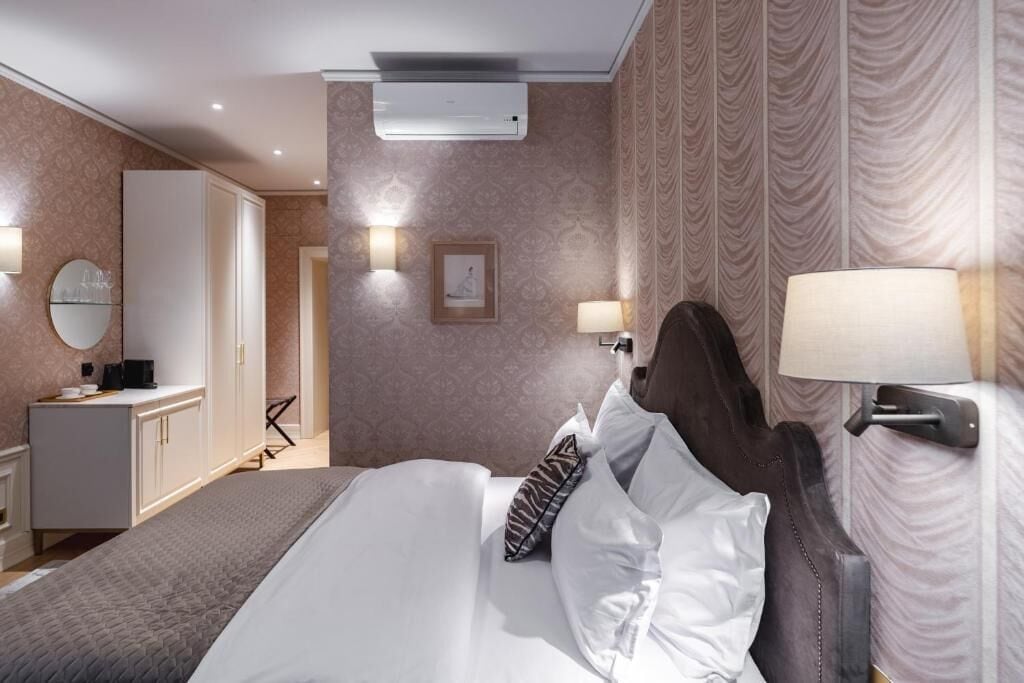 A hotel room with a bed, bedside lamps, a wardrobe, and an air conditioning unit.