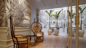 Una habitación amplia con un gran ventanal, una silla de mimbre, una mesita y plantas en macetas.