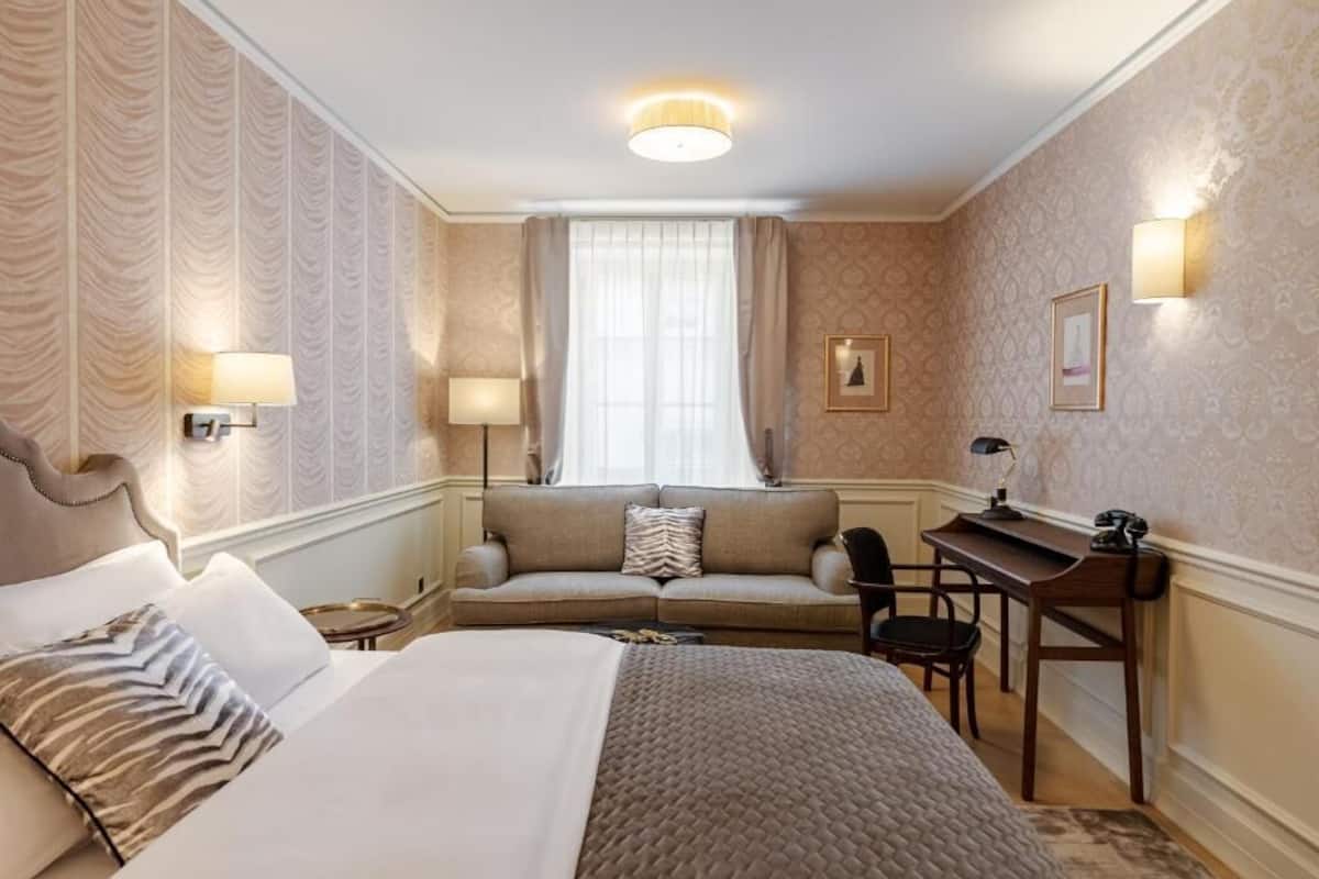 A hotel room with a bed, a sofa, a desk, and a chair. There are wall sconces and a window with curtains.