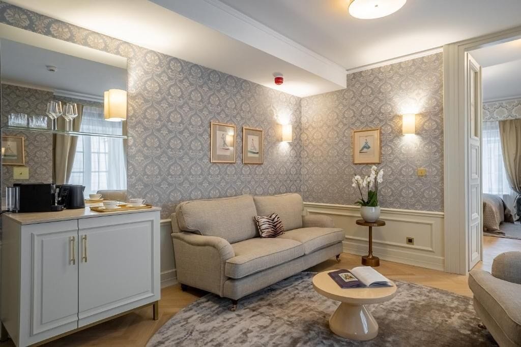 Ein Wohnzimmer mit Sofa, Couchtisch und Schrank. An den Wänden hängen Bilder und auf einem Beistelltisch steht eine Vase mit Blumen.