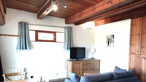 Uma sala de estar acolhedora com teto de madeira, uma televisão sobre um móvel de madeira, um sofá azul e uma mesa de jantar com cadeiras.