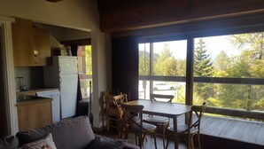 Uma sala de estar acolhedora com sofá, mesa de jantar com cadeiras, cozinha e grandes janelas que oferecem vista para árvores e um carro do lado de fora.