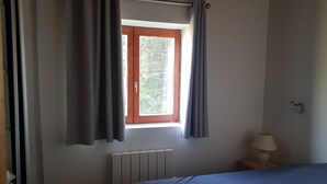 Quarto com cama, janela com cortinas, radiador e abajur sobre uma mesa de cabeceira.
