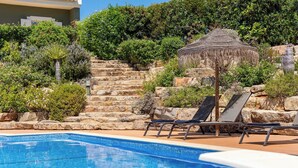 Piscina com espreguiçadeiras e um guarda-sol de palha, cercada por degraus de pedra e vegetação.