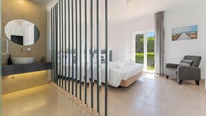 Quarto moderno com cama grande, área de estar, banheiro com pia e vista para o exterior.