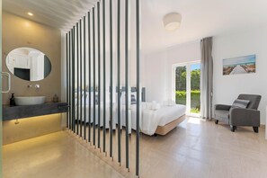 A modern bedroom with a large bed, a sitting area, a bathroom with a sink, and a view of the outdoors.