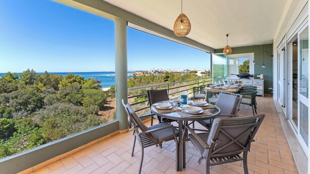 Varanda com mesa e cadeiras, oferecendo vista para o litoral com árvores e o mar.