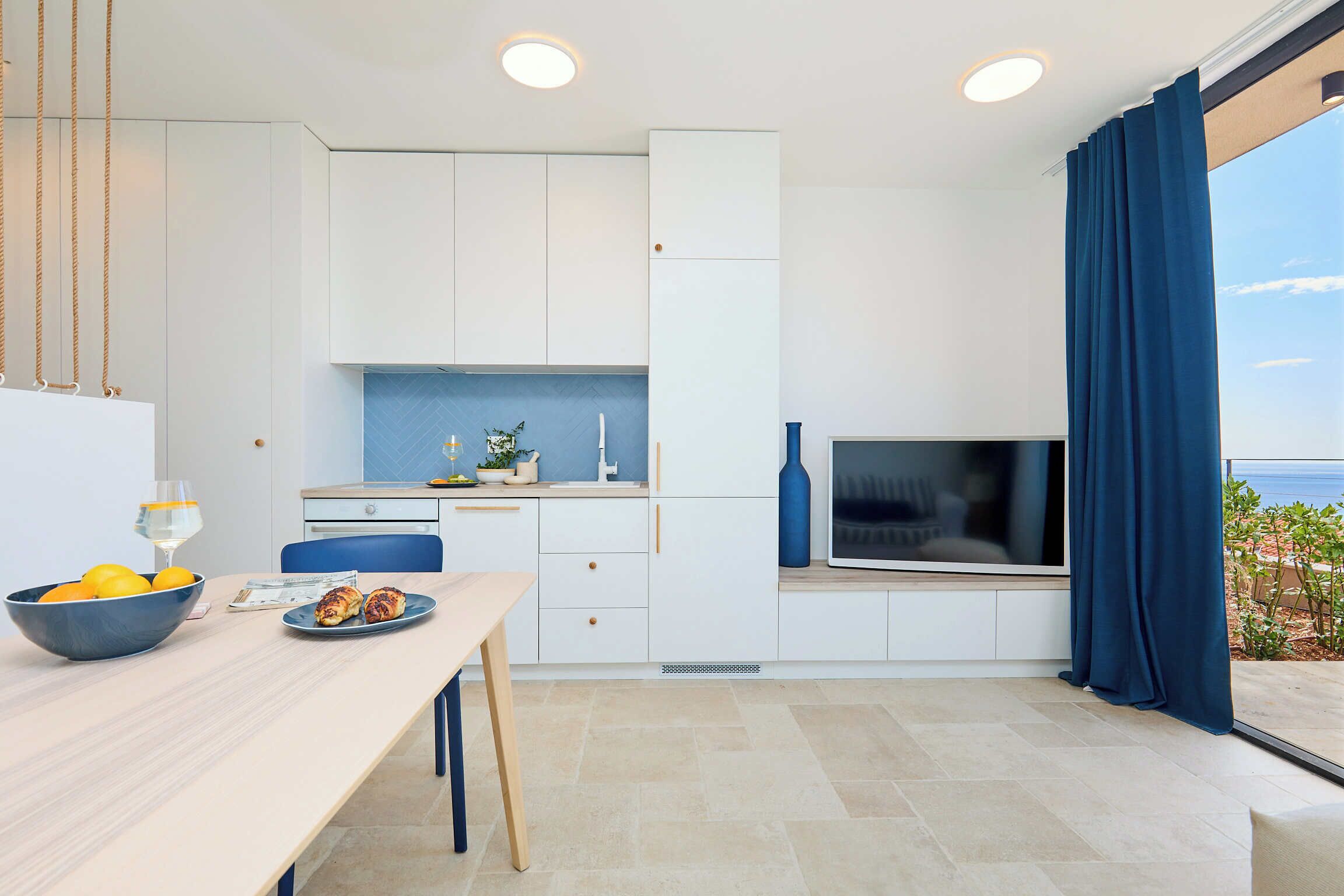 A modern kitchen with white cabinets, a dining table with a bowl of fruit, and a TV.
