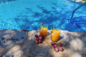 Dois copos de suco de laranja e um cacho de uvas sobre uma superfície de pedra perto de uma piscina.